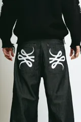 Pantalón de jean negro, corte baggy, confeccionado en denim crudo. Presenta un estampado serigrafiado en blanco con un diseño estilizado en ambos bolsillos traseros y un parche con logo en la cintura.