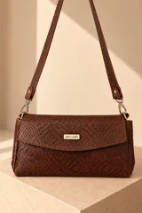 Cartera de cuero vacuno color marrón oscuro, con diseño trenzado y solapa frontal con placa metálica dorada grabada con la marca PECARÍ. Cuenta con correa larga para hombro y herrajes metálicos plateados.