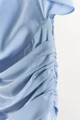Vestido midi de tejido satinado color celeste, con cuello barco y diseño asimétrico con mangas tipo capa. Presenta un detalle drapeado en la cintura y abertura en la espalda.