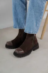 Bota corta de cuero gamuzado color marrón chocolate, con punta cuadrada y suela track. Presenta una correa con hebilla desmontable y cierre lateral con cremallera.