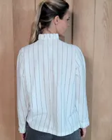 Camisa de algodón a rayas verticales blancas y azules, con cuello alto tipo gorguera con volados, cierre frontal de botones y mangas largas con puños con volados.