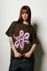 Remera de manga corta, corte regular fit, color marrón oscuro con un gran estampado frontal en rosa pálido que simula un logo estilizado o un símbolo abstracto.