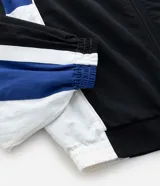Campera deportiva de poliamida con diseño de bloques de color en negro, blanco y violeta. Presenta cuello alto, cierre frontal con cremallera, puños y ruedo elásticos, y forro interior de poliéster.
