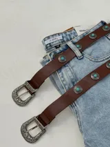 Cinto marrón de cuero con hebilla metálica labrada y apliques de piedras turquesas.