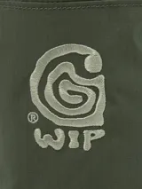 Bolso cilíndrico de diseño compacto en color verde militar, con cierre superior mediante cordón ajustable. Presenta un gráfico bordado en el frente y cuenta con una correa de hombro regulable y desmontable mediante ganchos metálicos.