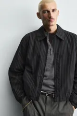 Cazadora de hombre de ajuste cropped fit, confeccionada en tejido de algodón con patrón de rayas verticales finas en tonos oscuros (negro/azul marino y marrón/beige). Presenta cuello solapa, cierre frontal de cremallera, bolsillos laterales y elástico en puños y bajo.