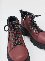 Botas de cuero color marrón oscuro con suela gruesa y dentada de goma negra, cordones estilo montaña en blanco y negro, y detalle de gamuza en el talón.