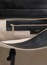 Bolso tipo shopper de gran tamaño confeccionado en tejido de canvas color beige. Cuenta con dos asas de hombro largas en contraste color negro, cierre de cremallera y un charm colgante decorativo en color negro.