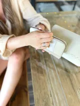 Billetera grande con cierre metálico, color beige claro, con paneles texturizados y un parche de cuero marrón con la marca escrita en cursiva.