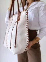 Cartera tipo tote de cuero vacuno con acabado texturizado en tono blanco hueso. Presenta detalles de costuras artesanales con tientos de cuero marrón en los bordes y correas de hombro a juego.