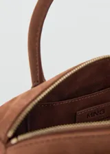 Bolso de mano tipo bowling de cuero gamuzado color marrón, con dos asas cortas y detalle de tiras con nudo y flecos colgantes en un lateral. Cierre superior con cremallera.