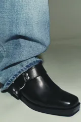 Zueco negro de cuero con punta cuadrada y taco bajo. Presenta una correa con tachas metálicas y una argolla metálica decorativa alrededor del empeine. Cuenta con un recorte en el talón.