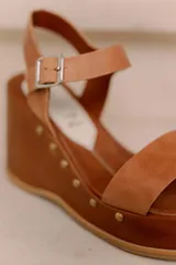 Sandalias de plataforma y taco alto confeccionadas en cuero vacuno marrón, con tira ancha en el empeine, tira ajustable con hebilla en el tobillo y detalles de tachas decorativas en la base.