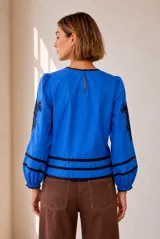 Blusa blanca de manga larga con detalles de bordados florales en tono grisáceo y bandas decorativas en el frente y mangas. Cuenta con cuello redondo y puños elásticos.