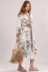 Túnica midi color blanco de manga larga, con escote en V profundo y lazo ajustable en la cintura. Presenta un estampado de paisaje desértico con palmeras y camellos, además de detalles geométricos en el escote, puños y cinturón.