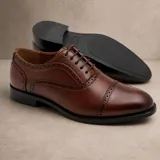 Zapato tipo Oxford de cuero sintético color marrón con detalles de perforaciones decorativas tipo brogue en la puntera y laterales. Cuenta con cordones al tono y suela de goma negra antideslizante.