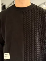 Sweater de punto oversize color marrón, con cuello redondo y diseño combinado: la mitad derecha cuenta con tejido de ochos y la izquierda es lisa. Incluye una etiqueta decorativa blanca en el frente inferior derecho.