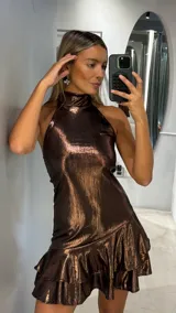 Vestido corto de cuello alto y estilo halter, confeccionado en tejido brillante con acabado metalizado en tono chocolate. Presenta un diseño ajustado en el torso con un ruedo de doble volado que aporta movimiento.