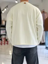 Sweater de punto liviano color beige, con textura marcada, cuello redondo y mangas largas de corte relajado, con pequeño estampado tipográfico negro en el lateral inferior.