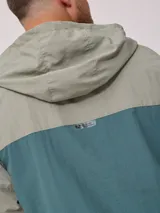 Campera rompevientos con cierre frontal, confeccionada en nylon con diseño de bloques de color en tonos verde azulado y gris claro. Cuenta con capucha, puños elásticos y cordón de ajuste en el ruedo.