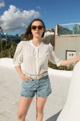 Camisa de estilo romántico color blanco, confeccionada en tejido liviano con textura de rayas verticales. Presenta mangas al codo con frunces en los hombros, cuello redondo y cierre frontal con botones. Incluye delicados detalles de puntilla de algodón en el pecho y en los bordes de las mangas.