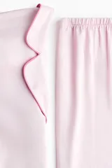 Pijama de dos piezas en tejido fluido de satén color rosado con ribetes en color contrastante. Camisa de manga larga con cuello abierto, botones adelante y bolsillo superior abierto, junto con un pantalón de pierna amplia.
