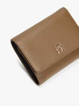 Billetera trifold de Tommy Hilfiger en color marrón, confeccionada en poliuretano con textura granulada. Presenta un diseño plegable con solapa frontal y el logo metálico TH en tono dorado en el centro.