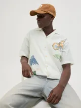 Camisa de algodón color crudo con estampado de flores y billetes, cuello resort, calce relajado, mangas cortas y cierre de botones.