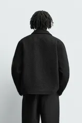 Sobrecamisa de corte relajado con textura de tejido estructurado en color gris y negro. Presenta cuello clásico, cierre frontal con botones, mangas largas con puños abotonados y bolsillos de plastrón en el pecho.