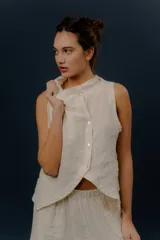 Chaleco de lino color marfil o beige claro, con diseño sin mangas, cuello alto amplio, cierre frontal con botones y bolsillos delanteros amplios. Presenta un corte asimétrico y relajado.