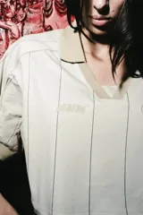 Camiseta crop de manga corta con cuello polo, confeccionada en tela técnica tipo dry fit. Presenta un diseño a rayas verticales finas en color beige, con paneles laterales de malla en tono camel y detalles de parches bordados en el frente.