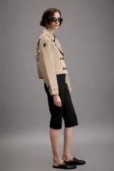 Trench corto de color beige con cuello de solapa, cierre frontal cruzado con botones y mangas largas con trabillas ajustables en los puños. Presenta un diseño estructurado con hombreras y bolsillos delanteros de vivo.