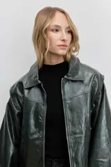 Cazadora de estilo bomber confeccionada en material efecto piel color verde oscuro. Presenta cuello camisero, cierre frontal con cremallera, mangas largas abullonadas con puños elásticos y un corte ligeramente cropped.