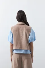 Chaleco de pana color marrón oscuro, con cuello camisero, cierre frontal de botones metálicos dorados y dos bolsillos frontales con detalle de bordado geométrico.