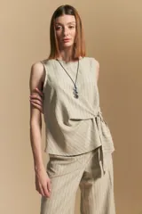 Top sin mangas color beige con finas rayas verticales negras. Presenta un diseño con lazada ajustable en el lateral de la cintura.