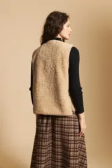Chaleco de textura tipo corderito en color beige, con cierre frontal de botones a presión y bordes ribeteados. Presenta un diseño sin mangas con silueta relajada.