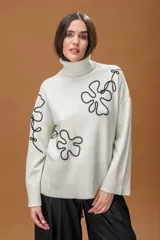 Sweater de punto color crudo con cuello alto, diseño holgado y hombros caídos. Presenta apliques de líneas orgánicas en relieve de color negro que recorren el frente y las mangas, con terminaciones acanaladas en puños, cuello y cintura.