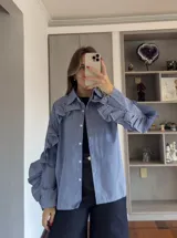 Camisa de corte recto con estampado de cuadros vichy en tonos azul y blanco. Presenta cuello clásico, cierre frontal con botones y volados decorativos que recorren la parte superior de las mangas y los hombros.