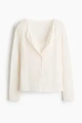 Saco entallado de punto suave, color blanco, con cuello redondo y cierre frontal de botones. Incorpora un panel interior cosido con efecto de top lencero con escote en V y ribete de encaje, creando un diseño de superposición.