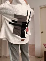 Campera de corderito blanco con capucha, de corte holgado y diseño tipo collage en la espalda con parches geométricos en tonos gris, negro y detalles en color. Incluye etiqueta de marca en la parte inferior trasera.