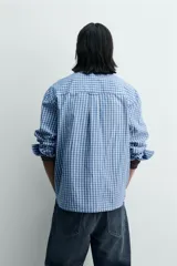 Camisa de manga larga con cuello solapa y bolsillo plastrón en el pecho. Presenta un estampado de cuadros pequeños en tonos azul y blanco. Cierre frontal con botones a presión.