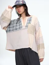 Blusa de estilo patchwork con diseño de cuello polo y escote en V. Combina diferentes texturas y tejidos en tonos neutros, incluyendo paneles de tela lisa, texturizada y a cuadros. Presenta mangas largas amplias con puños y un corte holgado.