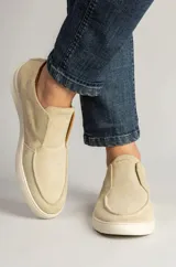 Zapatillas de gamuza color arena, diseño sin cordones estilo slip-on, con costuras decorativas en el empeine y suela de goma blanca.