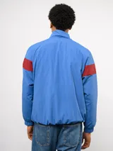 Buzo deportivo de nylon con medio cierre frontal y cuello alto. Presenta un diseño de bloques de color en azul, blanco y bordó, con el escudo del Club Nacional de Football y el logo de Umbro en el pecho.