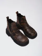 Botas de caña baja con punta cuadrada, confeccionadas en una combinación de cuero liso y gamuza en tonos marrones. Presentan un diseño con múltiples cortes y costuras visibles, cierre elástico lateral y suela de goma dentada tipo track.