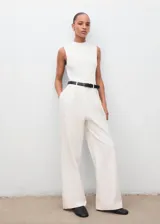 Mono largo de diseño recto y estilo minimalista, color blanco. Presenta cuello perkins, sin mangas con tirantes anchos, detalle de pinzas delanteras y cierre de cremallera en la parte posterior. Incluye bolsillos laterales y un cinturón negro con hebilla metálica.