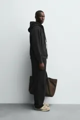 Sudadera con capucha y cierre de cremallera, color negro, de corte relajado y tejido de algodón compacto y pesado con interior rizo. Posee bolsillos frontales tipo canguro y acabados en rib.