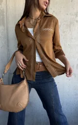Camisa de gamuza color camel, con cuello clásico, cierre frontal de botones y detalle de bordado geométrico en color crema a lo largo de la botonadura y en el bolsillo frontal. Presenta mangas largas con puños doblados.