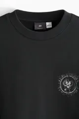 Buzo negro de algodón de peso medio con cuello redondo, hombros caídos y remates acanalados. Presenta un estampado frontal de una calavera rodeada de texto y texto en ambas mangas en tipografía gótica.