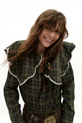 Camisa de manga larga confeccionada en popelina de algodón con estampado de cuadros en tonos verde oscuro y amarillo. Presenta un cuello amplio con bordes ondulados y terminaciones en encaje de algodón crudo, cierre frontal con botones de nácar y lazo de ajuste en el cuello.